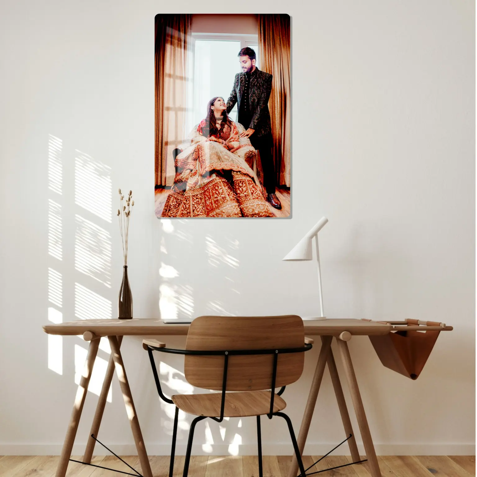 This acrylic print showcases a couple in traditional Indian wedding attire. The bride, seated, wears a richly embroidered red and gold lehenga, while the groom stands beside her, dressed in a black sherwani. The warm sunlight filters through a window, framed by golden curtains, creating a romantic and elegant atmosphere. The acrylic print is mounted above a minimalist desk, adding a personal and artistic touch to the room.