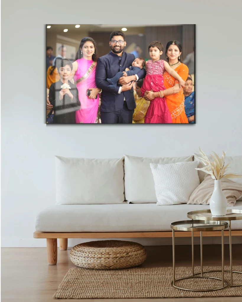 A simple, neutral-toned living room with a white sofa and a wicker stool. Above the sofa is a large acrylic photo print featuring a family portrait. The group, dressed in vibrant traditional attire, includes a man in a navy blue suit, women in orange and pink outfits, and several children, capturing a joyful family gathering.