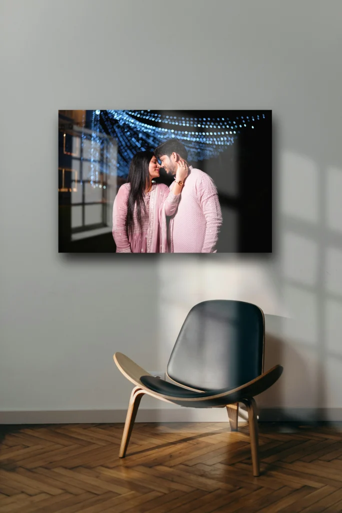 A modern minimalist space with a wooden floor and a single chair in front of an acrylic photo print on the wall. The photo features a couple in matching pink outfits sharing a romantic moment under blue-lit strings of lights, creating an intimate and dreamy atmosphere.