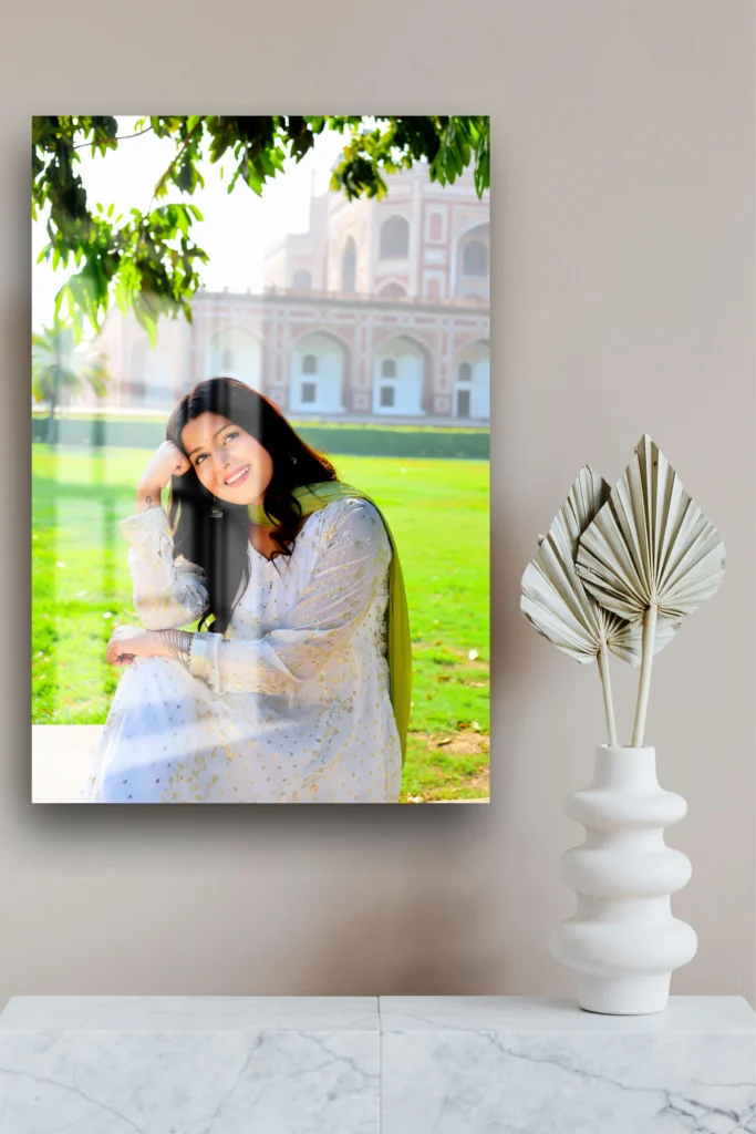 This acrylic print features a woman sitting outdoors in front of a historical monument, smiling gracefully. She is dressed in a light-colored traditional outfit with gold accents. The setting is lush and green, with tree branches overhead casting soft shadows. The acrylic print, displayed on a neutral-colored wall alongside modern white vases, exudes a calm and peaceful vibe, perfect for enhancing a space with both elegance and warmth.